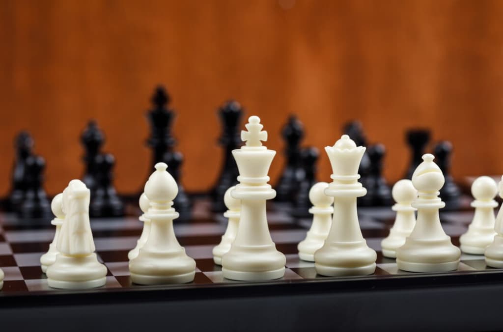 White chess pieces on a board, with black pieces slightly blurred in the background