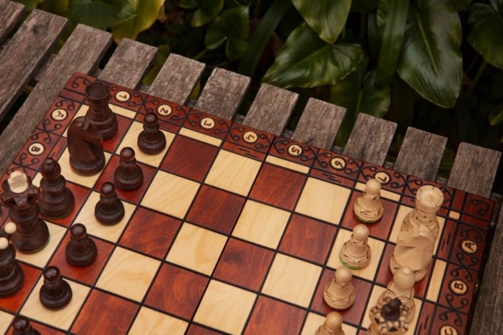 Chessboard on a wooden table, with dark and light pieces set against a backdrop of lush greenery