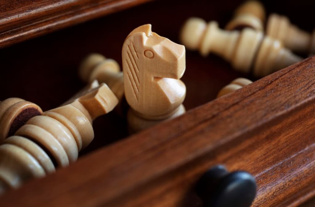 A carved wooden knight stands among fallen chess pieces in a rich, mahogany drawer